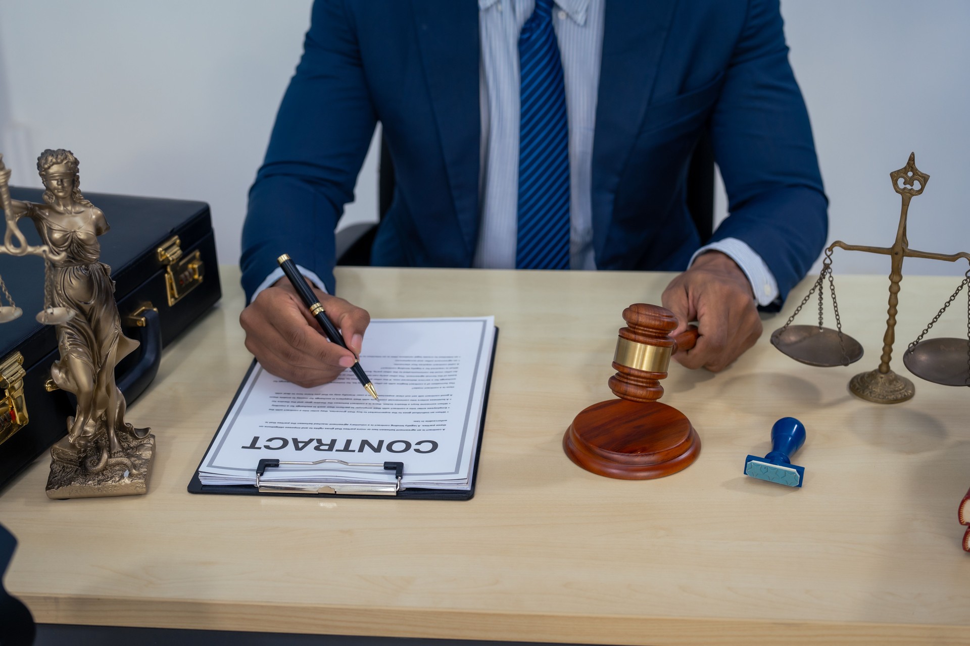 An Asian businessman in a formal suit works alone at a lawyer's desk, by golden scales, law books, leather bags, and a Lady Justice statue, emphasizing personal freedom and legal empowerment.